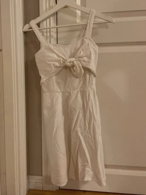 Vit kort klänning från Hollister - Supersöt vit klänning från Hollister med knyt framtill och breda axelband. Klänningen är kort och har en enkel, somrig design i mjuk bomull. Perfekt för varma dagar och enkel att styla med sneakers eller sandaler.
