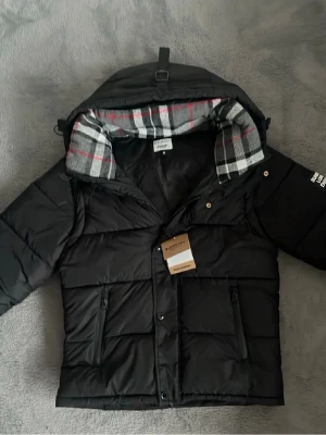 Svart dunjacka  - Svart dunjacka f med avtagbar huva och rutigt foder i grått, vitt och rött. Jackan har dragkedja, två fickor med dragkedja framtill och en modern, boxig passform. Perfekt för kalla dagar och enkel att matcha med olika outfits.