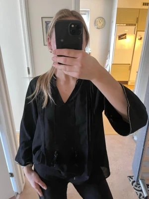 Svart blus med snörning - Svart blus med v-ringning, dekorativ snörning med tofsar och tunna veck framtill. Blusen har trekvartsärm och en lätt, luftig känsla. 