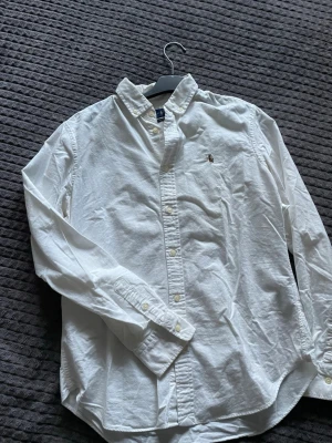 Vit skjorta från Ralph Lauren - Klassisk vit skjorta från Ralph Lauren med button-down krage och broderad logga på bröstet. Skjortan har lång ärm, normal passform och är tillverkad i mjuk bomull. Perfekt till jeans eller chinos för en clean och stilren look.