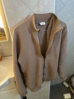 Beige ziptröja från John Henric - Stilren beige ziptröja från John Henric med diskret rutstruktur i materialet. Tröjan har ribbade muddar och krage samt dragkedja framtill. Perfekt för lager-på-lager och enkel att matcha med andra plagg.