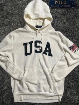 Vit hoodie från Polo Ralph Lauren - Vit hoodie från Polo Ralph Lauren med stor svart 'USA'-text på bröstet och amerikansk flagga med 'POLO' på vänster ärm. Klassisk känguruficka framtill och justerbar huva med dragsko. Tillverkad i mjukt bomullsmaterial för skön känsla.