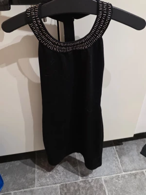 Svart kort klänning med pärldetaljer - Snygg svart kort klänning med rund halsringning dekorerad med silvriga pärlor. Ärmlös modell som ger en elegant och stilren look. Perfekt för dig som vill sticka ut med detaljerade accessoarer på klänningen.