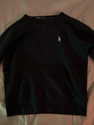 Svart tröja från Polo Ralph Lauren - Klassisk svart tröja från Polo Ralph Lauren med rund halsringning och broderad logga på bröstet. Tröjan har långa ärmar och ribbade muddar vid ärmslut och nederkant. Perfekt för en stilren och enkel look. Stlk 10-12