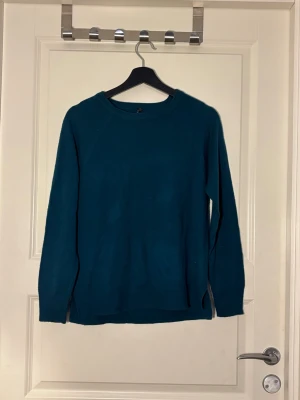 Mörk turkos stickad tröja - En enkel och stilren mörk turkos stickad tröja med rund halsringning och långa ärmar. Tröjan har en rak passform och ribbstickade muddar vid ärmslut och nederkant. Perfekt att matcha med jeans eller kjol.