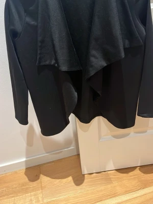 Svart omlott cardiganjacka - Snygg svart cardiganjacka med omlott-design och långa ärmar. Jackan har en mjuk och följsam känsla, perfekt att slänga på sig över en t-shirt eller topp. Stilren och enkel att matcha med det mesta i garderoben.