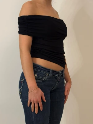Svart offshoulder topp - svart offshoulder topp med åtsittande passform och rynkade detaljer vid ärmarna. Toppen är croppad och har en stilren, enkel design som passar perfekt till jeans eller kjol. Tillverkad i mjukt stretchigt material för en bekväm känsla.
