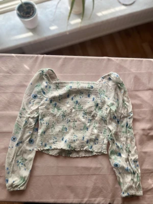 Blommig blus med puffärm från KappAhl - Supersöt blommig blus från KappAhl med smockad kropp och långa puffärmar i transparent tyg. Toppen har en fyrkantig urringning och är dekorerad med små blå och gröna blommor på vit botten. Perfekt för en trendig och romantisk look.