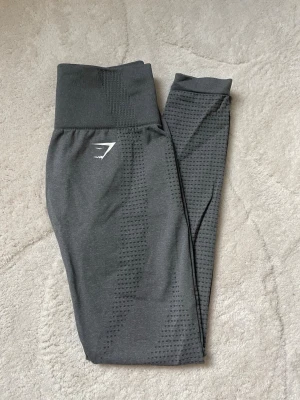 Träningsbyxor Gymshark - Snygga mörkgrå leggings från Gymshark med hög midja och diskret mönster längs benen. Stretchigt material som sitter skönt och följsamt. Perfekta för träning eller chill. Gymshark-logga framtill.