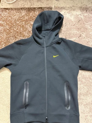 Svart Nocta Nike Tech Tracksuit - Snygg svart hoodie och byxor från Nike med gul broderad Swoosh på bröstet. Dragkedja framtill och två fickor med svarta dragkedjor. Tillverkad i mjukt och tjockt material, perfekt för chill eller träning. Klassisk huva för extra stil och komfort. 