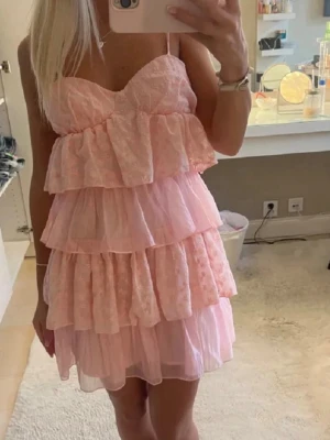 Rosa volangklänning med spetsdetaljer - Supersöt rosa kortklänning med flera lager volanger och tunna axelband. Klänningen har spetsdetaljer och en luftig, romantisk look som passar perfekt till sommaren. Materialet känns lätt och flowy, vilket ger en feminin känsla. Klänningen passar som storlek S💖