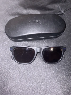 Svarta solglasögon från BOSS - Stilrena svarta solglasögon från BOSS med klassisk rektangulär form. Kommer med originalfodral i svart. Perfekta för att lyfta din look med en tidlös vibe. Köpta på room1006 i emporia, Malmö, finns kontoutdrag vid behov och intresse för bevis. 2600nypris solglasögon med tillhörande etui. Självklart äkta