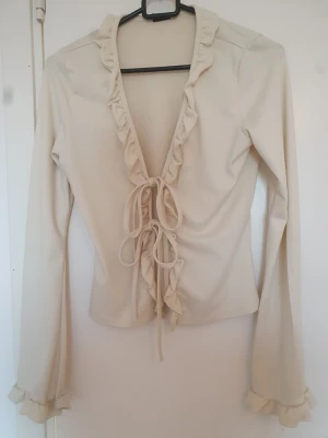 Beige omlottopp med volang och knyt - Supersöt beige omlottopp med volangkant längs urringningen och knyt framtill. Långärmad modell med lätt utsvängda ärmslut och volangdetaljer. Perfekt att styla med jeans eller kjol för en trendig look.