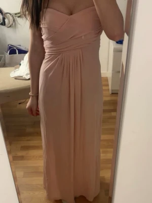 Ljusrosa långklänning från Mango - Säljer en elegant ljusrosa långklänning från Mango med omlottdetaljer upptill och axelbandslös design. Klänningen har ett mjukt, fallande tyg och markerad midja som ger en snygg siluett. Perfekt för speciella tillfällen!