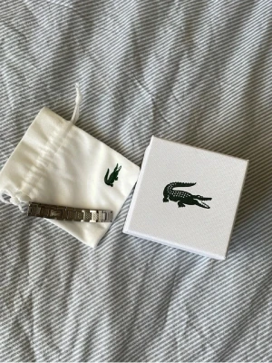 Silverfärgat armband från Lacoste - Snyggt armband i silverfärgad metall från Lacoste med diskret logotyp ingraverad på länkarna. Kommer med original ask, tygpåse och påse från Lacoste – perfekt för dig som gillar stilrena accessoarer.