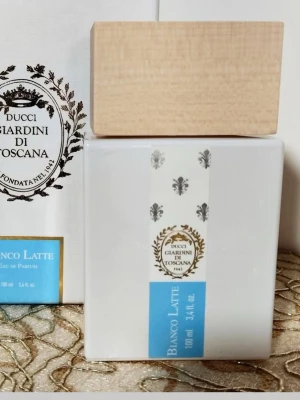 Giardini di Toscana Bianco Latte parfym - Säljer en Giardini di Toscana Bianco Latte Eau de Parfum 100 ml. Flaskan är vit med en ljusblå etikett och har en stilren, fyrkantig form. Korken är i ljust trä och ger en exklusiv känsla. Perfekt för dig som gillar unika dofter och snygg design på parfymflaskan.