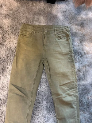 Olivgröna raka jeans - Säljer ett par olivgröna jeans med rak passform och klassisk femficksdesign. Jeansen har knapp och dragkedja framtill samt bälteshällor. Materialet är denim med en mjuk känsla och färgen är matt olivgrön, perfekt för en avslappnad stil.
