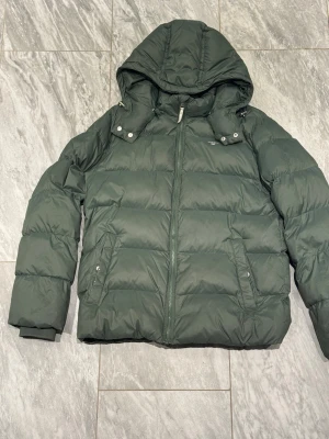 Mörkgrön pufferjacka med huva - Snygg mörkgrön pufferjacka med avtagbar huva och dragkedja framtill. Jackan har två sidofickor med tryckknappar och breda quiltade paneler för extra värme. Perfekt för kalla dagar och har en modern, boxig passform.