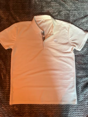 Vit pikétröja från Burberry - Klassisk vit pikétröja från Burberry med broderad logga på bröstet och typisk rutdetalj vid knäppningen. Tröjan har korta ärmar och mjukt bomullsmaterial som känns skönt mot huden. Perfekt till jeans eller shorts för en clean look.