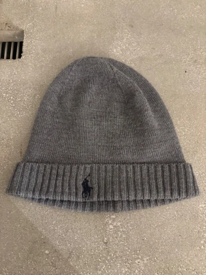 Grå mössa från Polo Ralph Lauren - (Använd max 1 gång inomhus!)Snygg grå mössa i mjuk och tvättbar merinoull från Polo Ralph Lauren. Klassisk ribbad kant och diskret broderad logga framtill. Perfekt accessoar för kalla dagar och enkel att matcha med olika stilar. (Ny pris 547)
