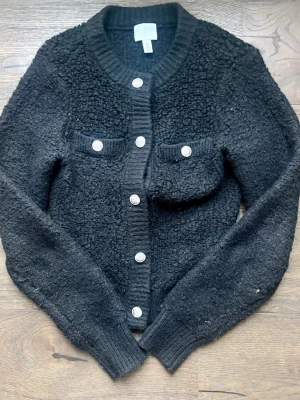 Svart kofta med knappar och fickor - Mysig svart kofta med strukturstickat material och silvriga knappar framtill. Två bröstfickor med lock och knappdetaljer. Långärmad och ribbstickade muddar vid ärmslut och nederkant. 