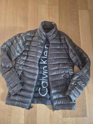 Grå dunjacka från Calvin Klein - Snygg grå dunjacka från Calvin Klein med avtagbar huva och tillhörande förvaringspåse. Jackan har quiltad design, hög krage och dragkedja framtill. Perfekt för kalla vinterdagar och enkel att packa ner när du är på språng.