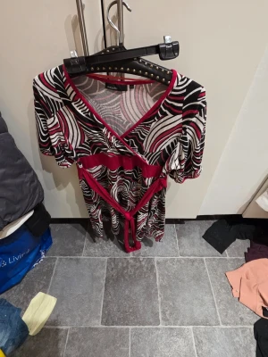 Mönstrad omlottklänning från KappAhl - Säljer en färgstark omlottklänning från KappAhl med svart, vitt och rött abstrakt mönster. Klänningen har korta puffärmar, markerad midja med knytband och V-ringning. Materialet är mjukt och stretchigt, perfekt för en trendig look.