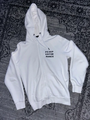 Vit hoodie från Peak Performance - Vit hoodie från Peak Performance med broderad logga på bröstet. Tröjan har huva med snörning, dragkedja framtill och två fickor. Tillverkad i mjuk bomullsmix, perfekt för chill dagar. Klassisk och clean look med ribbade muddar vid ärmslut och nederkant.