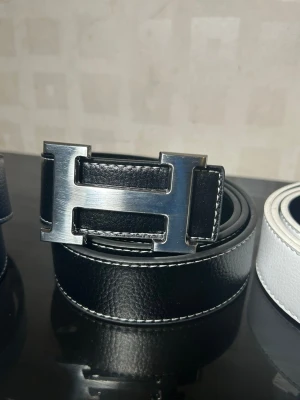 Svart bälte med H-spänne - Svart bälte med klassiskt H-spänne i silverfärgad metall. Bältet är tillverkat i äkta skinn med vit kontrastsöm längs kanterna. Stilren och tidlös design som lyfter vilken outfit som helst.