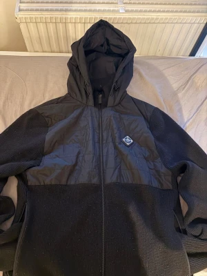 Svart hybridjacka från J.Lindeberg - Svart jacka från J.Lindeberg med huva och dragkedja. Jackan har stickade partier på ärmar och nederdel, samt quiltad polyester på bröst och rygg. Märkespatch på bröstet och praktiska fickor med dragkedja. Snygg och funktionell för kyliga dagar.