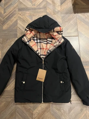 Burberry vändbar pufferjacka svart/rutig - Snygg vändbar pufferjacka från Burberry med klassiskt rutigt mönster på ena sidan och svart på andra. Jackan har huva, dragkedja, fickor med tryckknappar och metallogo på ärmen. Perfekt för dig som vill ha både stil och funktion.
