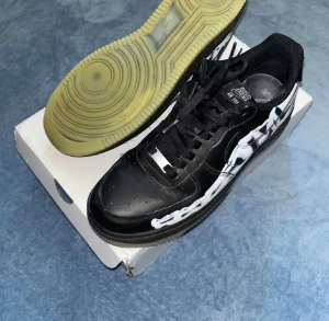 Nike Air Force 1 Skeleton svart/vit - Nike Air Force 1 Skeleton i svart skinn med vit skelett-design längs sidorna. Klassisk rund tå, snörning och platt sula med genomskinlig gummisula. Snygg och unik sneaker som sticker ut med sitt coola motiv och detaljer. Original låda och kvitto finns,köpta för 7999kr