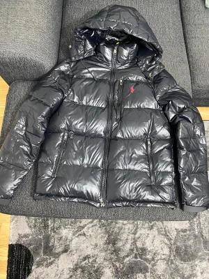 Svart pufferjacka från Polo Ralph Lauren - Svart pufferjacka från Polo Ralph Lauren med glansig finish, huva och flera dragkedjeförsedda fickor. Jackan har det klassiska röda Polo-logot på bröstet och quiltad design. Perfekt för kalla dagar med sin tjocka och fluffiga look. Säljer den var inte min stil har haft den runt 1 månad 