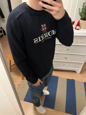 Mörkblå stickad tröja Morris - Snygg mörkblå stickad tröja från Morris med broderad logga och Union Jack på bröstet. Tröjan har rund hals, ribbade muddar och vit text framtill. Perfekt för dig som gillar klassisk stil med en twist. Storlek L men sitter som en M