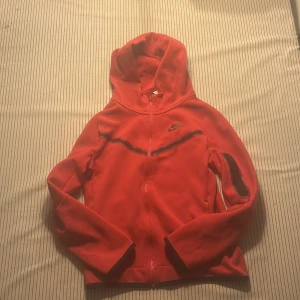 Röd hoodie från Nike med dragkedja - Nike hoodie i röd färg med svart logga på bröstet och svarta detaljer längs dragkedjan och ärmen. Tröjan har huva, hel dragkedja och långa ärmar. Perfekt för dig som gillar sportig och chill stil.