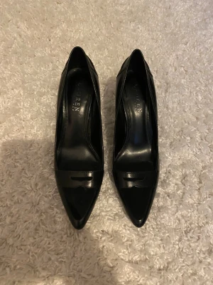 Svarta pumps från Lauren Ralph Lauren - Snygga svarta pumps från Lauren Ralph Lauren med spetsig tå och låg klack. Skorna är tillverkade i skinn och har en stilren design med dekorativ detalj framtill. Perfekta för dig som gillar klassisk och elegant stil.