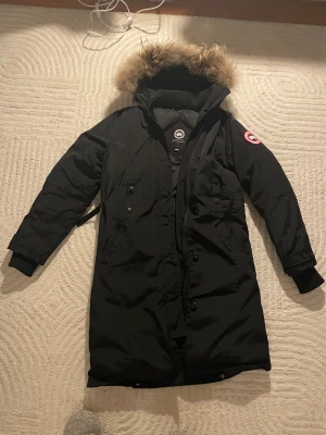 Canada Goose Kensington - Säljer en svart parkas från Canada Goose med ikonisk röd-vit-blå patch på ärmen och fluffig beige päls runt huvan. Jackan har dragkedja, knappar och ribbade muddar vid ärmslut. Perfekt för kalla vinterdagar och riktigt snygg streetstil.