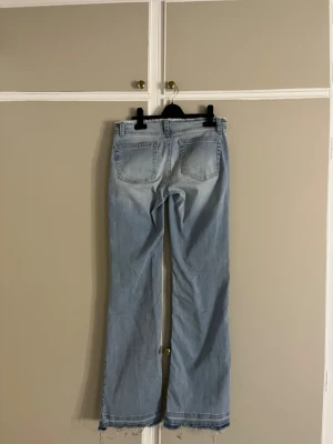 Ljusblå bootcut jeans från Nelly - Snygga ljusblå jeans från Nelly med bootcut passform och råa, fransiga kanter både i midjan och vid bensluten. Klassisk femficksmodell med gul kontrastsöm och knappstängning. Perfekt för dig som gillar en avslappnad och trendig look.