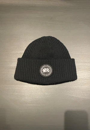 Svart mössa från Canada Goose - Svart mössa från CG med ribbad struktur och rund patch framtill med Arctic Program-logga. Klassisk uppvikt kant och mjukt material som håller dig varm. Perfekt för dig som vill ha en stilren look under kalla dagar.