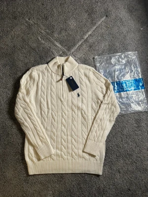 Vit stickad tröja Polo Ralph Lauren - Säljer en vit kabelstickad tröja från Polo Ralph Lauren i storlek S. Tröjan har half zip, ribbad krage och muddar, samt det klassiska broderade Polo-logot på bröstet. Materialet är mjuk ull och passformen är normal. Perfekt för kyliga dagar.