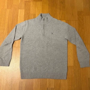 Grå stickad half zip-tröja Gant - Snygg grå stickad tröja från Gant med half zip och hög krage. Tröjan har ribbade muddar och liten broderad logga på bröstet. Perfekt för lager-på-lager och passar till både jeans och chinos. Pris kan diskuteras!