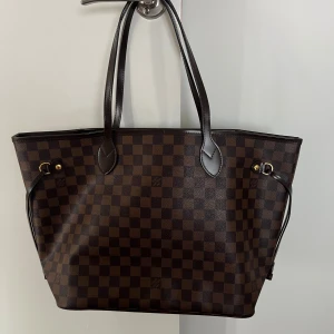 Neverfull väska - Snygg och rymlig Neverfull-väska i klassiskt brunt Damier Ebene-mönster. Väskan har mörkbruna handtag, guldfärgade metalldetaljer och rödrandigt innerfoder med dragkedjefack. Perfekt för dig som vill ha både stil och funktion. Väskan är i nyskick och bara använts några gånger. Inget slitage 