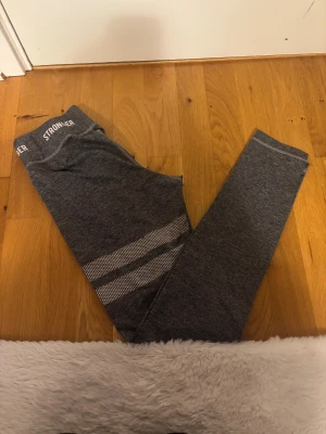 grå leggings från Stronger - Snygga grå leggings från Stronger med vita ränder i mesh på ena benet och logga i midjan. Stretchigt material som sitter skönt och passar perfekt till träning. Stilren design med smal passform och hög midja.