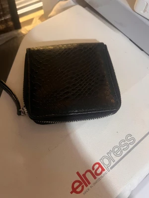 Svart plånbok i skinn med reptilmönster - Snygg svart plånbok i skinn med reptilmönster och dragkedja runtom. Insidan har flera kortfack och ett myntfack med knapp. Perfekt storlek för att ha med sig överallt och ger en cool vibe med sitt mönster.