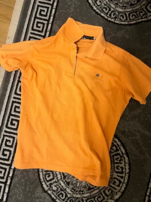 Orange pikétröja från Burberry - Säljer en orange pikétröja från Burberry med klassisk krage och knäppning framtill. Tröjan har Burberrys ikoniska rutiga detalj på insidan av kragen och en liten broderad logga på bröstet. Perfekt för dig som gillar färg och stil.
