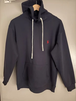 Mörkblå hoodie från Polo Ralph Lauren - Snygg mörkblå hoodie från Polo Ralph Lauren med vit snörning och klassisk röd logga på bröstet. Tröjan har huva, magficka och långa ärmar. Perfekt för dig som gillar stilrena och tidlösa plagg med premiumkänsla.ps. Aldrig använd, bara stått i garderoben.