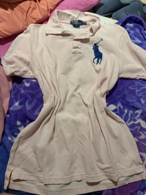 Ljusrosa pikétröja från Ralph Lauren - Ljusrosa pikétröja från Ralph Lauren med klassisk krage, korta ärmar och stor blå logga broderad på bröstet. Snygg detalj med blå siffra 3 på ena ärmen. Tillverkad i bomull för en skön och luftig känsla.