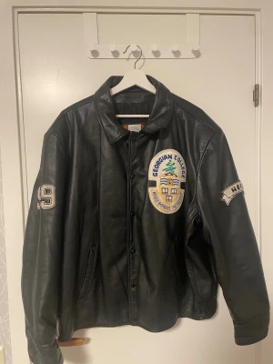 Svart skinnjacka Georgian College - Svart skinnjacka med broderade patches från Georgian College, Nursing. Jackan har klassisk krage, knappar framtill och två sidofickor. Fodret är quiltat och insidan har etikett med tillverkarinfo. Snygga detaljer som broderad text och siffror på ärmen.