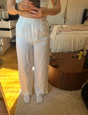 Vita linnebyxor med hög midja - Vta linnebyxor med hög midja och raka, vida ben. Byxorna har klassiska fickor bak och fram, bälteshällor och stängs med dragkedja och knapp.