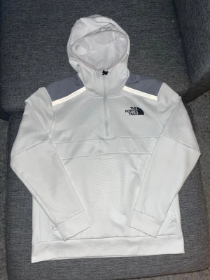 Vit hoodie från The North Face - Snygg vit hoodie från The North Face med grå detaljer på axlarna och reflekterande band. Tröjan har half zip, huva och långa ärmar. Materialet är polyester med en strukturerad yta som känns sportig och modern. Perfekt för dig som gillar streetwear och outdoor-vibe.